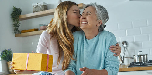 Jeune femme offrant un cadeau à sa mamie pour la fête des grands-mères dans une cuisine en lui faisant un bisou sur la joue.