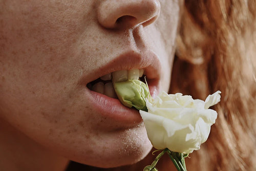 Bouche de femme rousse mordant une rose blanche dans une ambiance sensuelle pour illustrer les aphrodisiaques naturels.
