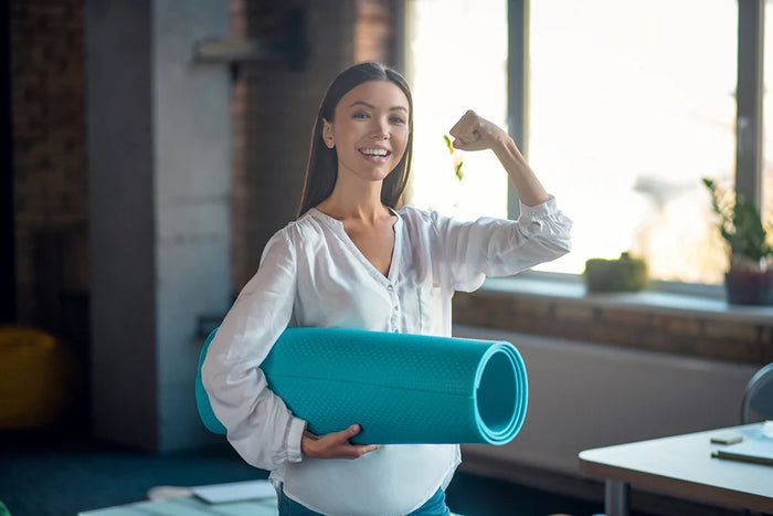 Femme souriante tenant un tapis de yoga le poing levé pour illustrer le fait de retrouver la forme